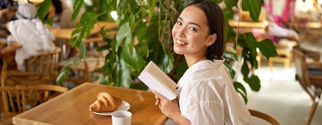 Бесплатное фото Красивая молодая азиатка с книгой в руках сидит в кафе, пьет кофе и ест