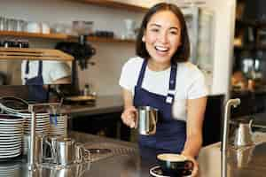 Free photo beautiful young female barista making cappuccino pouring steamed milk for latte art into cup give yo