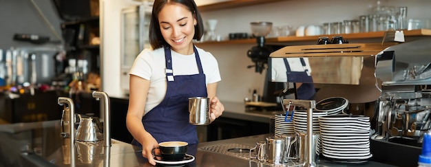 Free photo beautiful young female barista making cappuccino pouring steamed milk for latte art into cup give