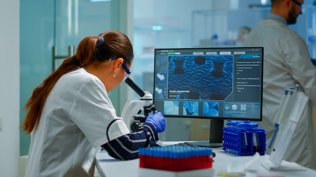 Free photo biotechnology scientist researching in laboratory using microscope and typing on pc. chemist examining virus evolution using high tech for scientific research of vaccine development against covid19
