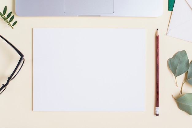 Free photo blank white paper; pencil; eyeglasses; leaves and laptop on beige backdrop