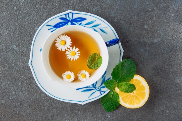 Brewed chamomile tea with half of lemon and green leaves in a cup and saucer on grey stucco background, top view.