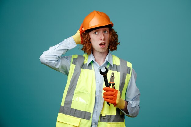 Builder young man in construction uniform and safety helmet holding wrench and pliers looking confused with hand on his head for mistake standing over blue background