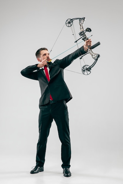 Free photo businessman aiming at target with bow and arrow, isolated on gray studio wall