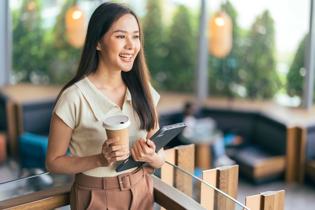 Foto gratuita programmatore femminile asiatico casual che codifica stile di vita di momad digitale tenere in mano tablet e tazza di caffè sorridente allegro sentimento pacifico workign ovunque in piedi al ristorante caffetteria che cambia stile di vita