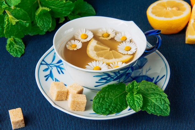 Free photo chamomile tea in a cup and lemon slices, brown sugar cubes and green leaves side view on a dark placemat background