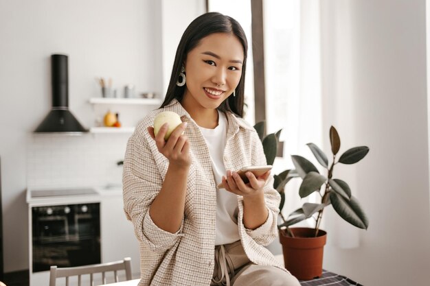 Free photo charming asian woman in beige cardigan and pants looks into camera smiles and holds phone attractive lady in stylish outfit sits on table and holds apple