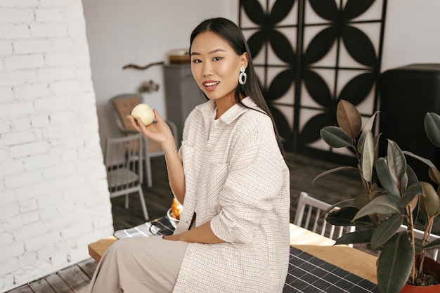 Free photo charming brunette tanned woman in beige cardigan and pants looks into camera smiles and holds apple pretty asian lady sits on wooden desk on kitchen