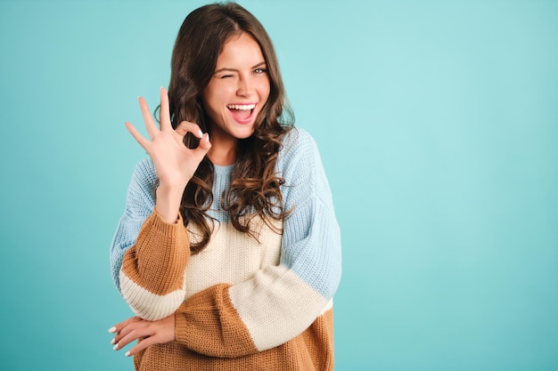 Foto gratuita ragazza allegra in un maglione accogliente che mostra felicemente il gesto giusto e strizza l'occhio a porte chiuse su sfondo blu