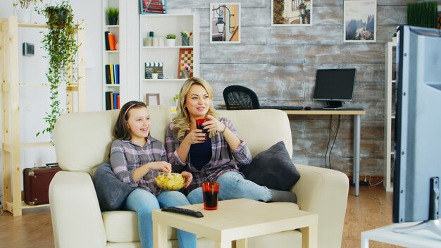 Cheerful mother and daughter sitting on the couch in living room watching tv, eating chips and drinking soda.