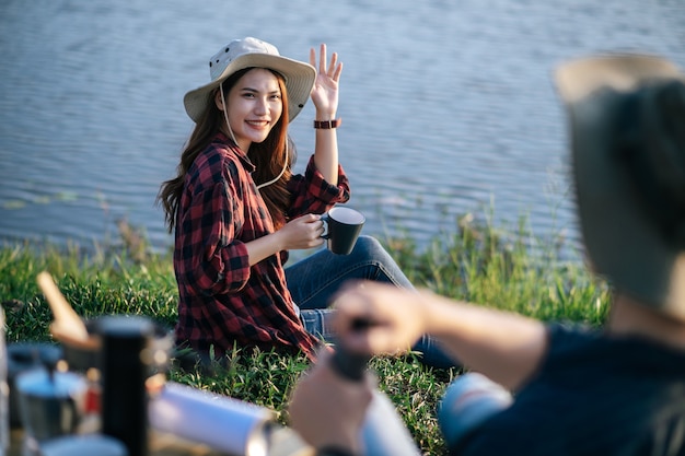 Free Photo cheerful young backpacker couple wearing trekking hat sitting near lake with coffee set and making fresh coffee grinder while camping trip on summer vacation