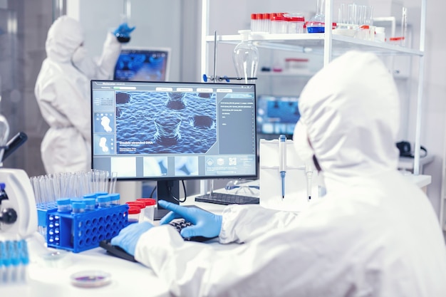 Free photo chemist sits at his workplace in laboratory analyze coronavirus on computer dressed in ppe