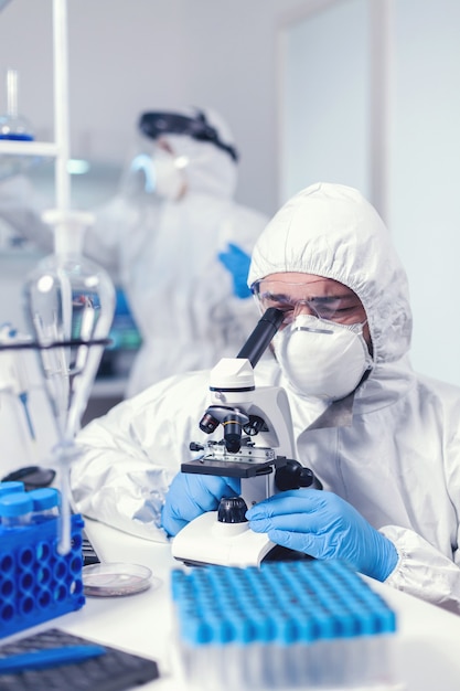 Free photo chemist with face mask and ppe developing vaccine for covid looking through microscope. scientist in protective suit sitting at workplace using modern medical technology during global epidemic.