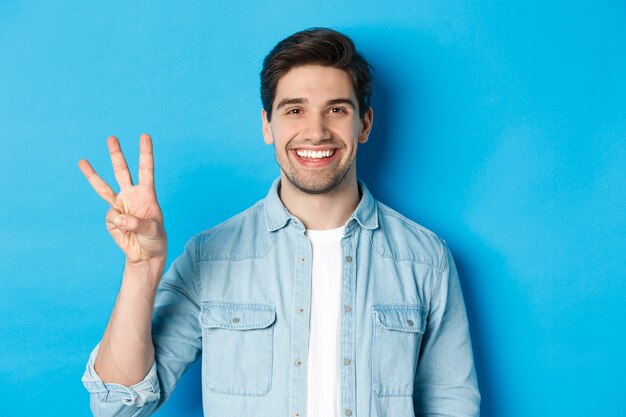 Close-up of handsome man smiling, showing fingers number three, standing over blue background