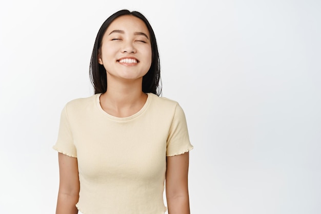 Free photo close up portrait of carefree asian woman smiling with closed eyes daydreaming imaging something standing in yellow tshirt over white background