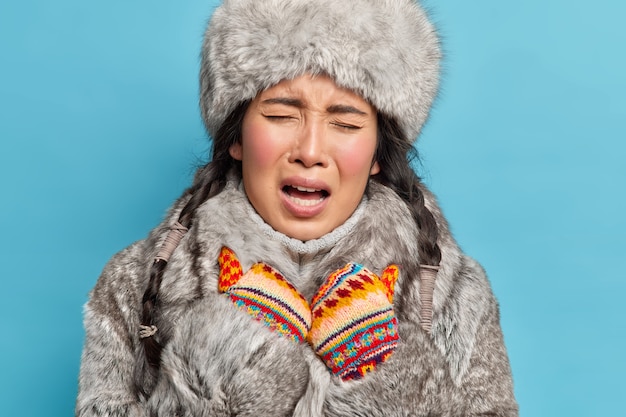 Free photo close up shot of displeased young asian inuit woman wears knitted mittens and winter outfit feels cold during frosty winter isolated over blue wall