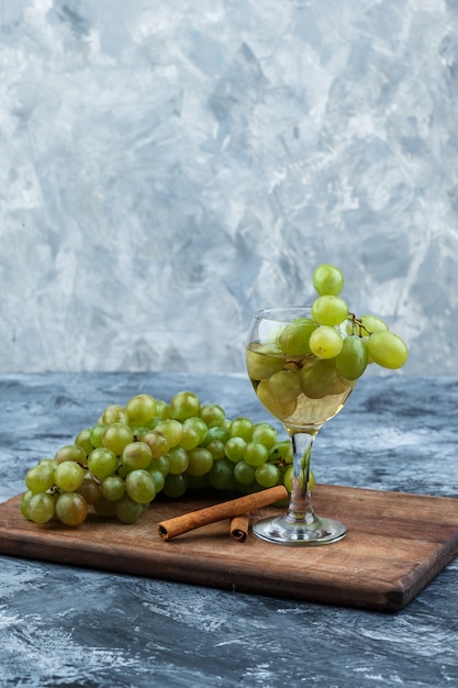 Free photo close-up white grapes, glass of whisky, cinnamon on cutting board on dark and light blue marble background. vertical