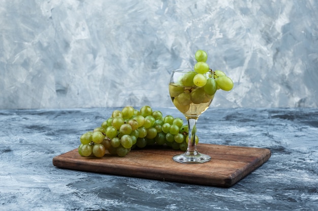 Free photo close-up white grapes, glass of whisky on cutting board on dark and light blue marble background. horizontal