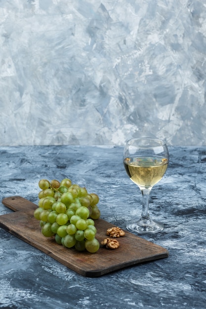 Free photo close-up white grapes, walnuts on cutting board with glass of whisky on dark and light blue marble background. vertical