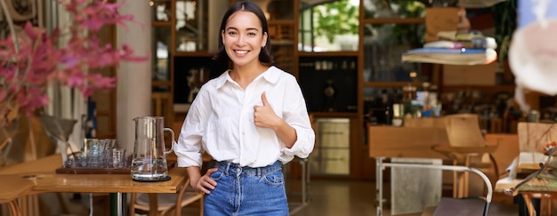 Free photo confident asian businesswoman showing thumbs up standing near entrance of her cafe or restaurant