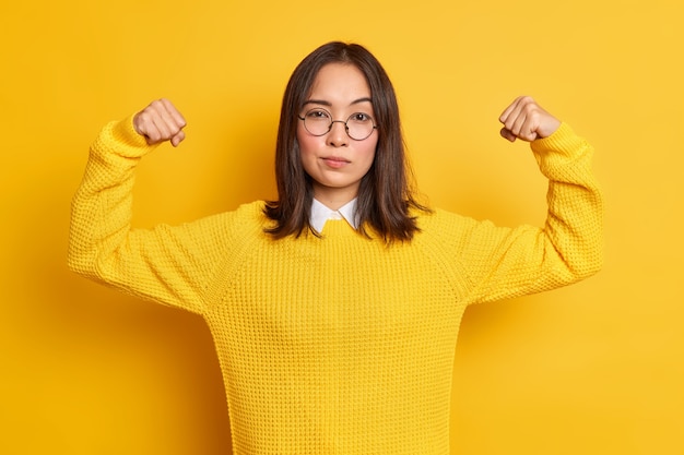 Free Photo confident young asian woman shows arm muscles feels like hero demonstrates her power and strength looks seriously wears round optical glasses sweater.