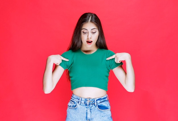 Free photo confident young girl standing and pointing at down side on red background.