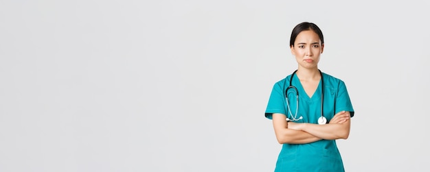 Free photo covid-19, healthcare workers, pandemic concept. skeptical and reluctant asian female doctor, tired nurse in scrubs cross arms and frowning, smirk displeased, standing white background.