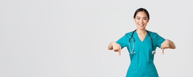 Free photo covid-19, healthcare workers, pandemic concept. smiling pleasant asian female doctor, therapist or physician in scrubs with stethoscope, pointing fingers down, show clinic banner