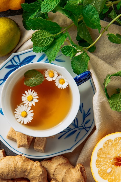 Free photo cup of chamomile tea with sliced lemon, ginger, brown sugar cubes and green leaves in a saucer on piece of cloth background, high angle view.