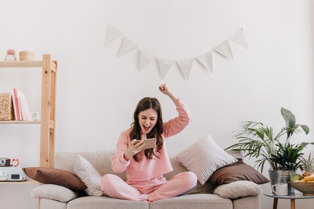 Curly woman in pink suit rejoices in winning game in smartphone