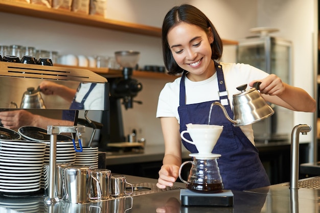 Free photo cute brunette girl barista cafe staff pouring water from kettle and brewing filter coffee behind cou