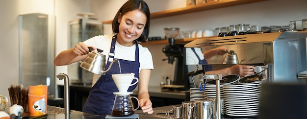 Free photo cute brunette girl barista cafe staff pouring water from kettle and brewing filter coffee behind