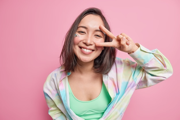 Free photo cute positive asian woman with dark hair shows v sign peace gesture looks happily  dressed in casual shirt enjoys good day isolated over pink wall expresses carefree emotions.