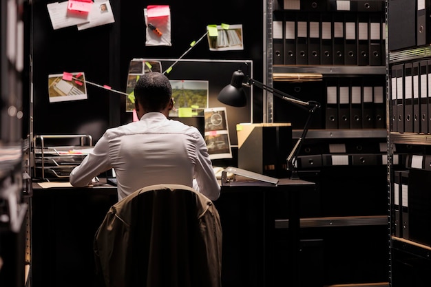 Free photo detective sitting at desk and analyzing evidence scheme hanging on wall. police law enforcement professional solving crime, studying clues and crime scene photos on board at night time