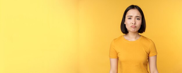 Disappointed asian girl sulking looking upset feel unair standing in yellow tshirt over white backgr