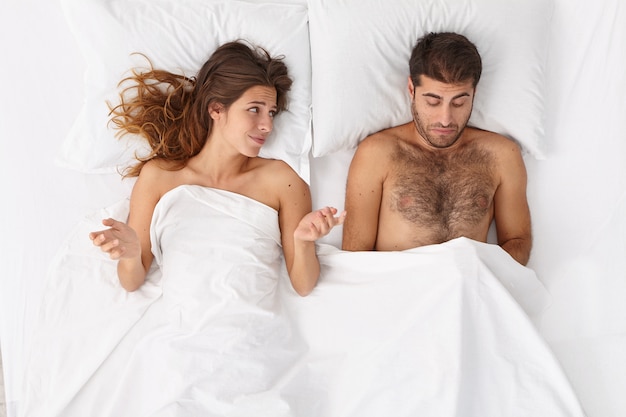 Free photo disappointed man has erectile dysfunction during sex, his woman partner lies near under white blanket, puzzled with husbands impotence, spreads hands sideways. sexual problems. men health concept