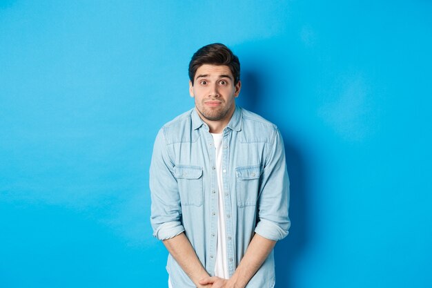 Embarrassed guy want to pee, waiting in line for toilet, standing against blue background.