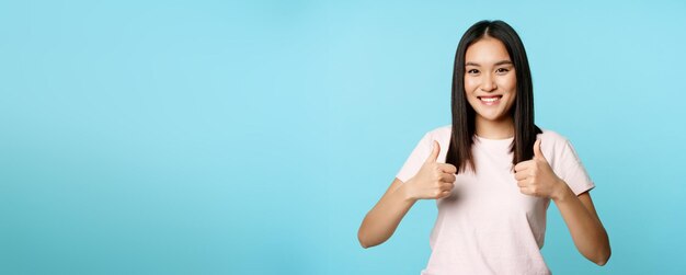 Foto gratuita la ragazza asiatica entusiasta che mostra i pollici in su gradisce e approva sorridente soddisfatto in piedi sul dorso blu