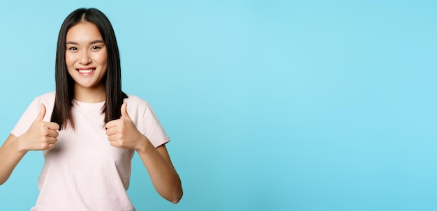 Foto gratuita la ragazza asiatica entusiasta che mostra i pollici in su gradisce e approva sorridente soddisfatto in piedi sul dorso blu