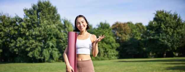 Free Photo excited asian girl in sportswear holds rubber mat for yoga looks surprised and happy stands in park