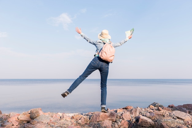 Free photo excited young woman standing on top of rock holding map in hand overlooking the sea against blue sky