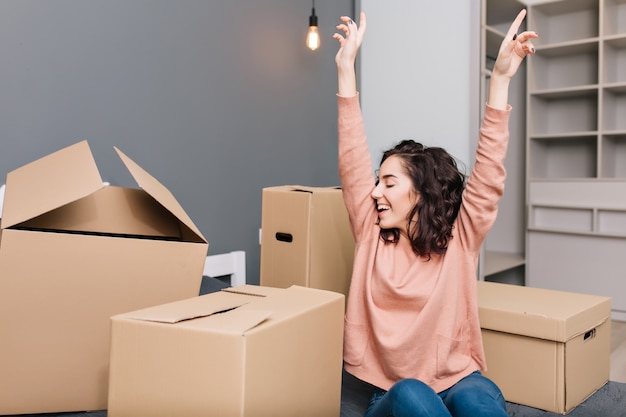 Free photo expressing brightful true emotions, positivity of young pretty woman with short brunette curly hair on bed surround carton boxes in modern apartment. enjoying moving, happiness at new home