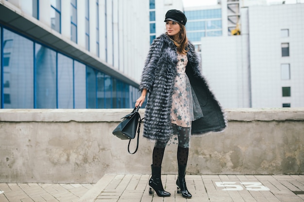 Free photo fashionable smiling woman walking in city in warm fur coat, winter season, cold weather, wearing black cap, dress, boots, holding leather bag, street fashion trend