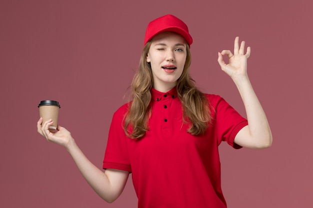 Free Photo female courier in red uniform holding coffee cup winking on pink, uniform service delivery job worker