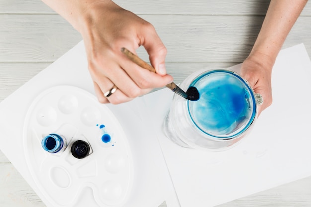 Free photo female hand painting on glass jar with paint brush over wooden desk