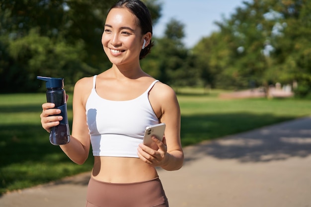 Free photo fitness woman with water bottle and smartphone jogging in park and smiling looking at her mobile pho