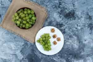 Free photo flat lay white grapes in bowl with grapes, walnuts in a white plate on dark blue marble background. horizontal