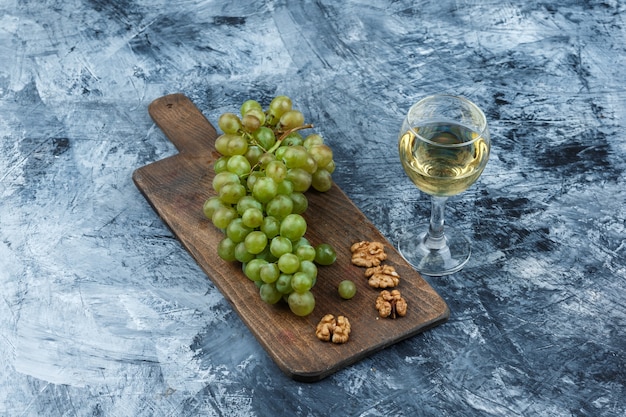 Free photo flat lay white grapes, walnuts on cutting board with glass of wine on dark blue marble background. horizontal