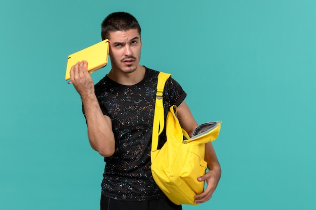 Front view of male student in dark t-shirt yellow backpack thinking on light blue wall