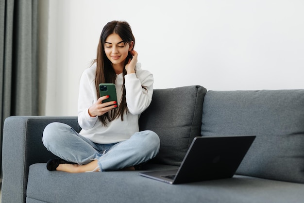 Full length portrait of carefree woman holding her mobile and communicating with smile and delight on couch at home